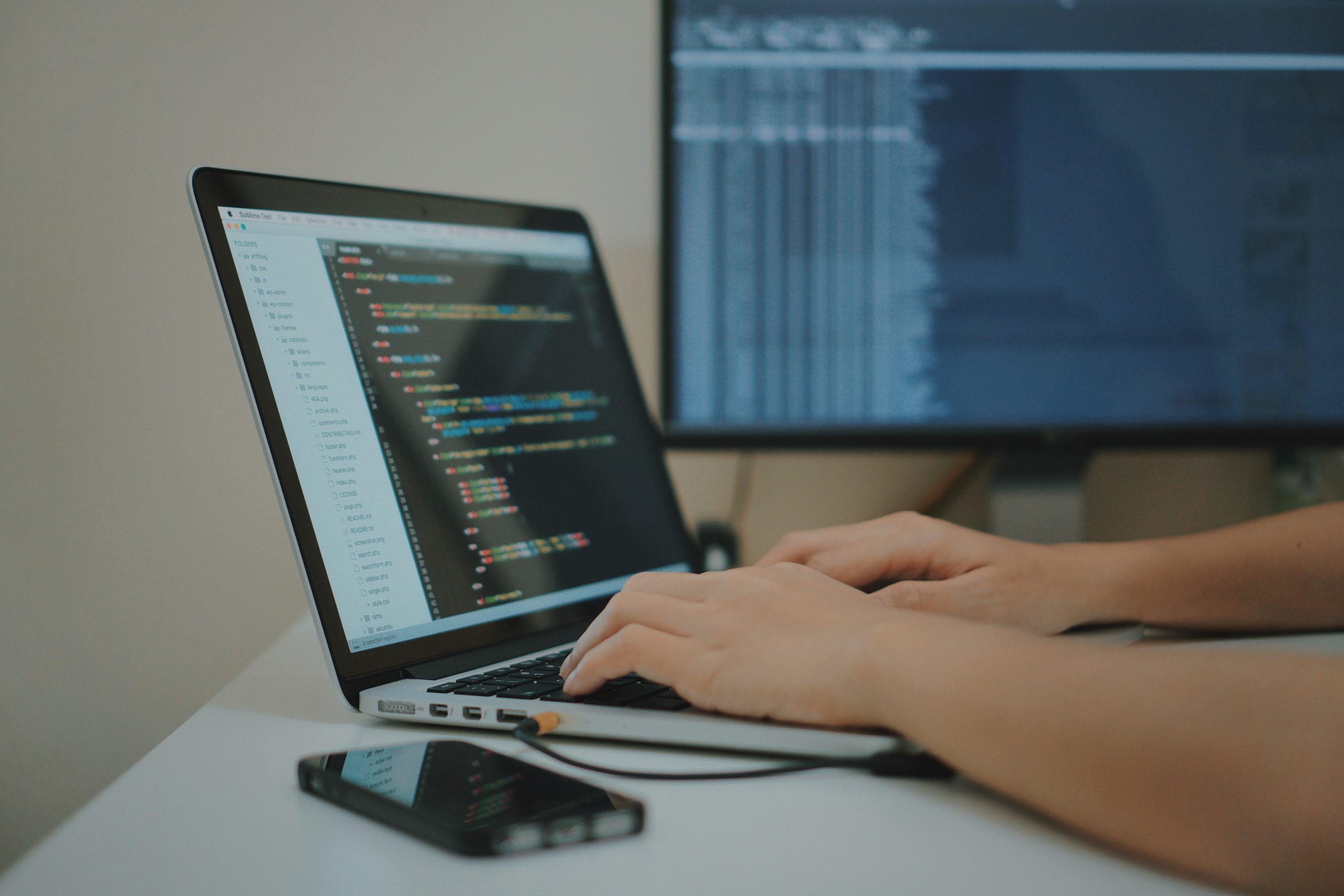 Person coding on a laptop with two screens and a smartphone nearby.